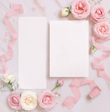 Paper cards between light pink roses and silk ribbons on pink top view,  wedding mockup. Romantic scene with vertical blank cards and pastel flowers flat lay. Valentines, Spring or Mothers day concept