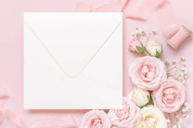 Square envelope between light pink roses and silk ribbons on pink top view,  wedding mockup. Romantic scene with blank envelope and pastel flowers flat lay. Valentines, Spring or Mothers day 