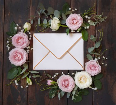 Envelope between pink and cream roses on brown wood top view,  wedding mockup. Romantic scene with envelope and pastel flowers flat lay. Valentines, Spring or Mothers day concept