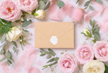 Sealed kraft paper envelope between light pink roses and silk ribbons on marble top view,  wedding mockup. Romantic scene with envelope and pastel flowers flat lay. Valentines, Spring or Mothers day
