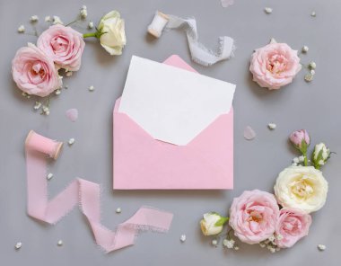 Card and envelope between light pink roses and silk ribbons on grey top view,  wedding mockup. Romantic scene with blank vertical card and pastel flowers flat lay. Valentines, Spring or Mothers day concept