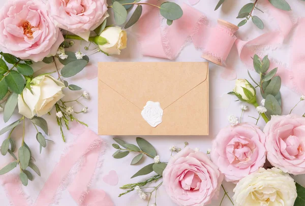 Sealed kraft paper envelope between light pink roses and silk ribbons on marble top view,  wedding mockup. Romantic scene with envelope and pastel flowers flat lay. Valentines, Spring or Mothers day