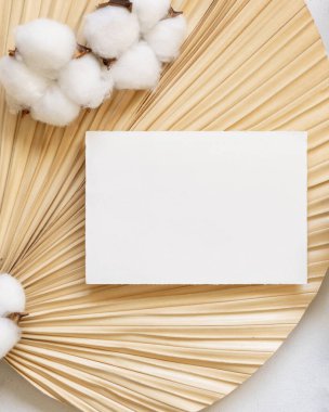 Blank card on dried palm leaf with cotton flowers top view, mockup. Romantic scene with horizontal paper card. Bohemian pastel greeting or wedding Invitation