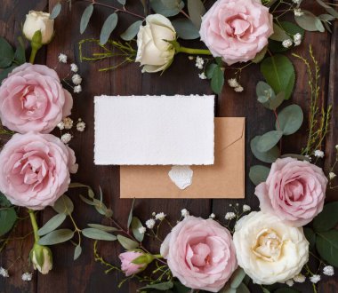 Card and envelope between pink and cream roses on brown wood top view,  wedding mockup. Romantic scene with cards, envelope and pastel flowers flat lay. Valentines, Spring or Mothers day concept