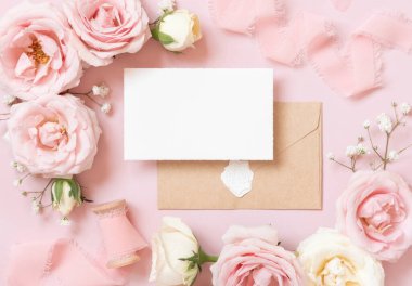 Card with envelope between light pink roses and silk ribbons on pink top view,  wedding mockup. Romantic scene with vertical blank card and pastel flowers flat lay. Valentines, Spring or Mothers day concept