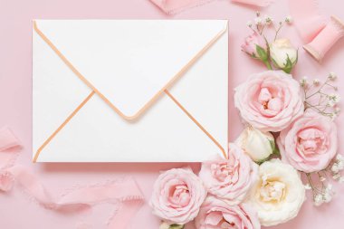 Envelope  between light pink roses and silk ribbons on pink top view,  wedding mockup. Romantic scene with blank envelope and pastel flowers flat lay. Valentines, Spring or Mothers day concept