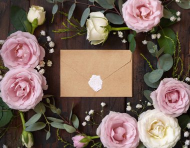 Sealed Envelope between pink and cream roses on brown wood top view,  wedding mockup. Romantic scene with envelope and pastel flowers flat lay. Valentines, Spring or Mothers day concept
