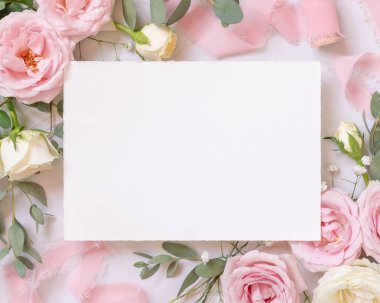 Blank card between light pink roses and silk ribbons on marble top view,  wedding mockup. Romantic scene with horizontal card and pastel flowers flat lay. Valentines, Spring or Mothers day