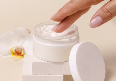 Açık kozmetik kavanozundan krem kullanan bir kadın eli, orkide çiçeklerinin yanında boş bir kapakla yakın plan, maket. Cilt bakımı ürün paketi, minimal bileşik