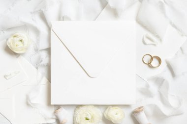 Krem güllerin yanındaki kare zarf, beyaz ipek kurdeleler, alyanslar üst manzara, düğün modeli. Pastel çiçeklerin olduğu romantik bir sahne. Sevgililer Günü, Bahar veya Anneler Günü konsepti