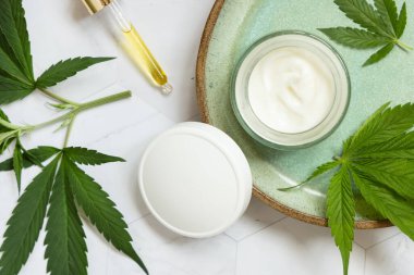 Yağlı boru ve beyaz kapaklı krema kavanozu beyaz mermer masa manzaralı tabakta yeşil kenevir yaprakları, maket. Organik cilt ürünleri, doğal kozmetik ürünleri.