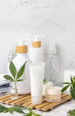 Yeşil kenevir yapraklarının yanındaki beyaz krem tüpü diğer şişelerin yanındaki banyoda, ahşap tepside, yakın plan, maket. Organik cilt ürünleri, doğal CBD kozmetik ürünleri