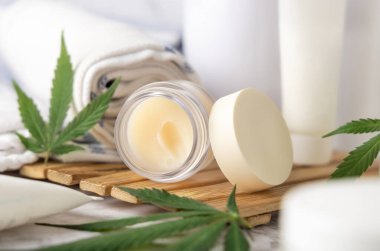 CBD dudak kremi ve yeşil kenevir yapraklı kavanoz şişelerin yanında ve banyoda havluyla ahşap bir tepsiye yakın. Organik cilt bakımı ürünü. Alternatif kozmetik