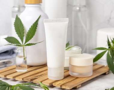 Yeşil kenevir yapraklarının yanındaki beyaz krem tüpü diğer şişelerin yanındaki banyoda, ahşap tepside, yakın plan, maket. Organik cilt ürünleri, doğal CBD kozmetik ürünleri
