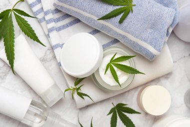 Havlunun üstünde yeşil kenevir yapraklarının yanında beyaz kapaklı krema kavanozu ve şişeler var. Organik cilt bakım ürünleri, alternatif CBD kozmetik ürünleri