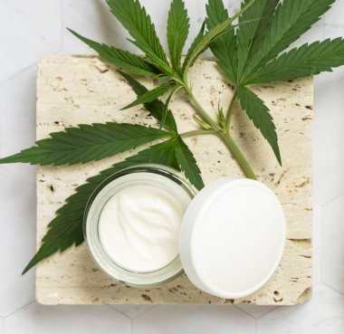 Açık krema kavanozu, boş kapaklı ve yeşil kenevir yaprakları bej taştan üst manzaralı. Organik çevre dostu CBD kozmetik ürünleri. Alternatif yaşam formu