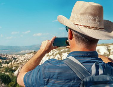 Tişörtlü, sırt çantalı ve kovboy şapkalı bir adam güneşli bir günde cep telefonuyla fotoğraf çekiyor. İnsanlar güzel manzarayı görmek için dağlarda yürürken durdular.