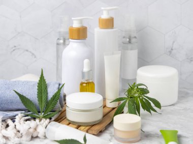 Yeşil kenevir yapraklarının yanındaki boş kozmetik kavanozları, tüpler ve şişeler diğer şişelerin yanındaki banyodaki ahşap tepside, yakın plan, maket. Organik cilt ürünleri, doğal CBD kozmetik ürünleri
