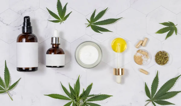 Cosmetic bottles, jar, pipette with CBD oil, capsules with hemp powder and infusion tea near green cannabis leaves on a marble table top view. Organic skincare and healthcare products. CBD Cosmetic