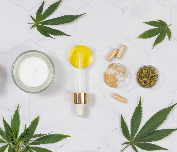 Cream jar, pipette with CBD oil, capsules with hemp powder and infusion tea near green cannabis leaves on a marble table top view. Organic skincare and healthcare products. CBD Cosmetic