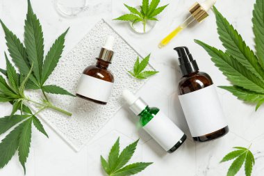Krema kavanozu, CBD yağlı pipet, yeşil kenevir yaprakları yakınında kenevir tozu ve infüzyon çayı bulunan kapsüller mermer masa manzaralı. Organik cilt bakımı ve sağlık ürünleri. CBD Kozmetik