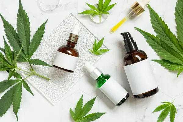 Cream jar, pipette with CBD oil, capsules with hemp powder and infusion tea near green cannabis leaves on a marble table top view. Organic skincare and healthcare products. CBD Cosmetic