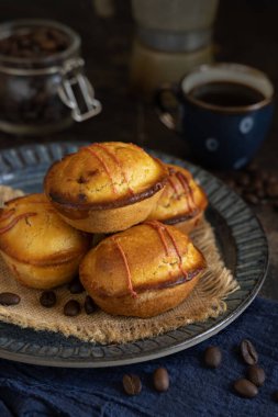 Kahve fincanının yanında koyu mavi bir tabakta Pasticciotto leccese hamur işi ve koyu masa üzerinde moka, tipik Apulian kahvaltısı, yakın çekim. Kahve çekirdekleri ve peçeteyle süslenmiş bir masa.