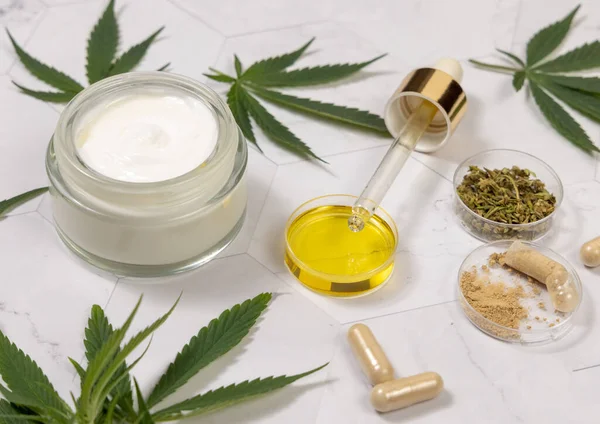 Cream jar, pipette with CBD oil, capsules with hemp powder and infusion tea near green cannabis leaves on a marble table close up. Organic skincare and healthcare products. CBD Cosmetic