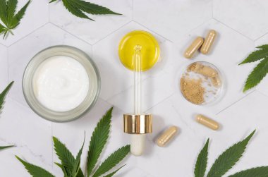 Kozmetik kavanoz, CBD yağlı pipet ve yeşil kenevir yapraklarının yakınındaki kenevir tozlu kapsüller mermer masa manzaralı. Organik cilt bakımı ve sağlık ürünleri. CBD Kozmetik