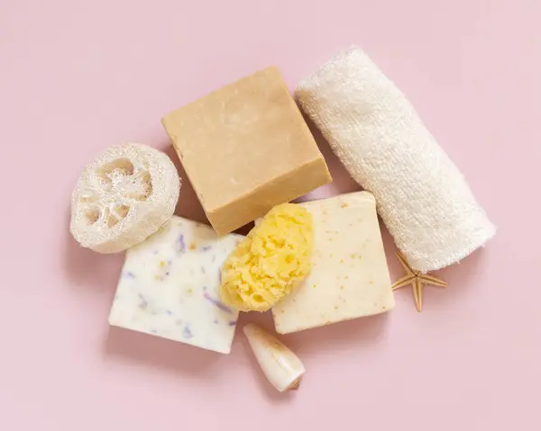 Beige soap bars, seashells, natural sponges and towel on light pink top view. Herbal products for face and body care. Using organic natural skincare product