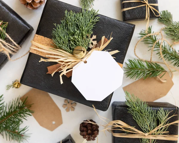 Black present with Christmas gift tag, fir tree branches, pine cones and holiday decorations, top view. Winter composition with blank label Mockup, copy space, top view