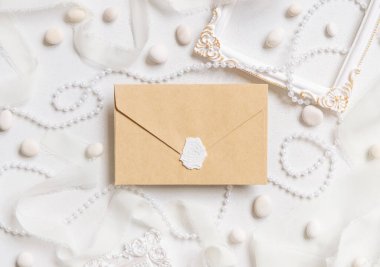 Beyaz çerçeveler, inciler, çakıl taşları ve beyaz masa manzaralı ipek kurdeleler yanında mühürlü bej bir zarf, düğün modeli. Boş zarfın olduğu romantik bir sahne. Sevgililer Günü, Bahar veya Anneler Günü konsepti