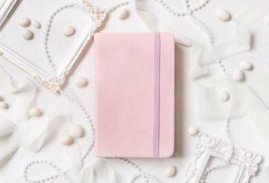 Çerçevelerin, incilerin, çakıl taşlarının ve beyaz masa manzaralı ipek kurdelelerin yanındaki açık pembe defter. Hardcover ders kitabının maketi olan romantik bir sahne. Sevgililer Günü, eğitici ve kız işi konsept