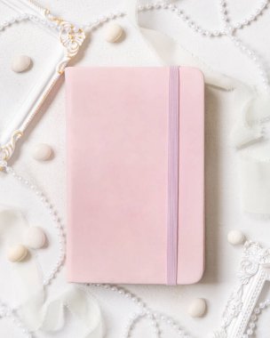 Çerçevelerin, incilerin, çakıl taşlarının ve beyaz masa manzaralı ipek kurdelelerin yanındaki açık pembe defter. Hardcover ders kitabının maketi olan romantik bir sahne. Sevgililer Günü, eğitici ve kız işi konsept