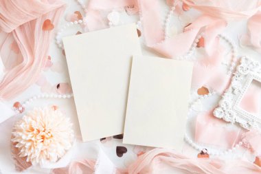 Kağıt kartlar pembe dekorasyonlar, kalpler ve beyaz masa manzaralı ipek kurdeleler, maket. Dikey boş kartların olduğu romantik bir sahne. Düğün, Sevgililer Günü, Bahar veya Anneler Günü kırtasiyesi