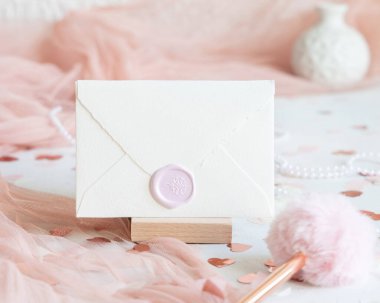 Pembe dekorasyonlar, kalpler ve beyaz masa üzerindeki ipek kurdeleler yakınındaki mühürlü zarf. Düğün, Sevgililer Günü, Bahar veya Anneler Günü kırtasiyesi için romantik bir model.