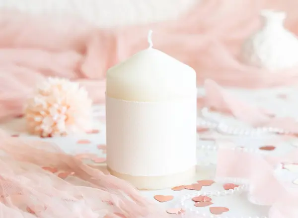 Candle with blank label near pink decorations, hearts and silk ribbons on white table close up. Romantic home decor