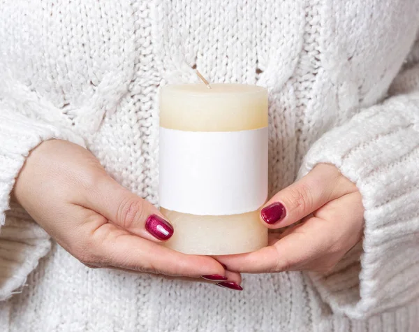 Woman in white sweater holding in hands cream candle with blank label, mock up, copy space, front view. Girl with red nails and pillar candle. Cozy holidays atmosphere