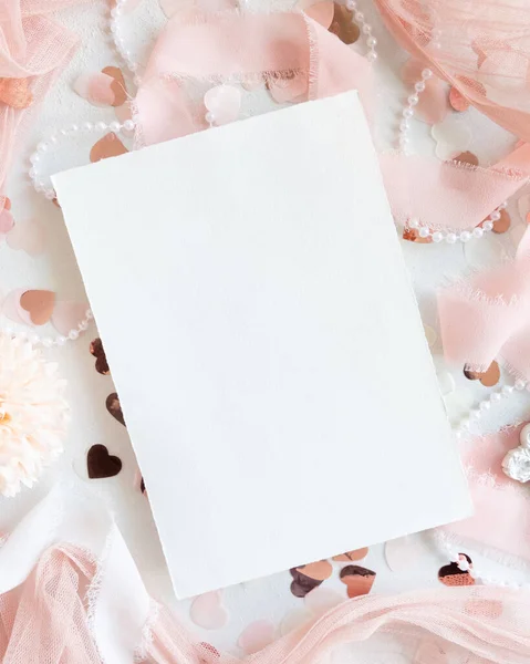 Blank paper card near pink decorations, hearts and silk ribbons on white table top view,  mockup. Romantic scene with vertical blank card flat lay. Wedding, Valentines, Spring or Mothers day stationery
