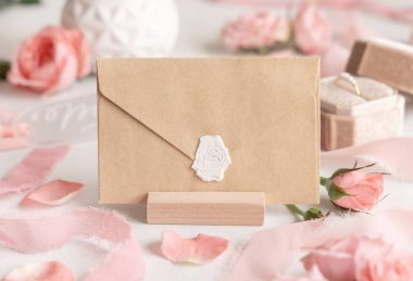 Açık pembe güller, taç yaprakları, bir kutuda nişan yüzüğü ve beyaz masadaki ipek kurdeleler yanında boş bir zarf. Zarf ve pastel dekorlu romantik bir sahne. 