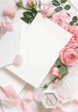Açık pembe güllerin yanındaki boş kart ve zarf, bir kutuda nişan yüzüğü ve beyaz masa manzaralı ipek kurdeleler, düğün modeli. Dikey kartı ve pastel dekoru olan romantik düz bir yatak. 