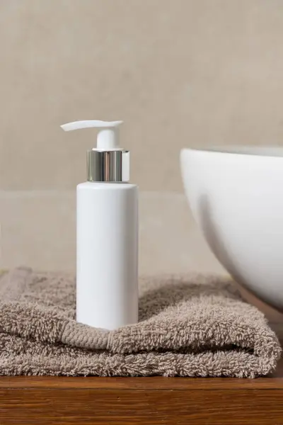 White one pump bottle near basin on folded towel on wooden countertop in beige bathroom, close up, cosmetic mockup.  Minimal composition for brand packaging