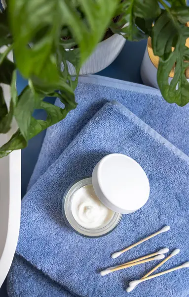 Opened cream jar with blank white lid and bamboo cotton swabs on blue towels near basin and green monstera blue countertop, top view, mockup.  Lifestile scene with skincare beauty product in bathroom
