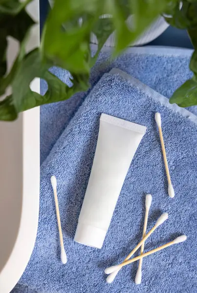 White cream tube and bamboo cotton swabs on blue towels near basin and green monstera on blue countertop, top view, mockup.  Lifestile scene with skincare beauty product in bathroom