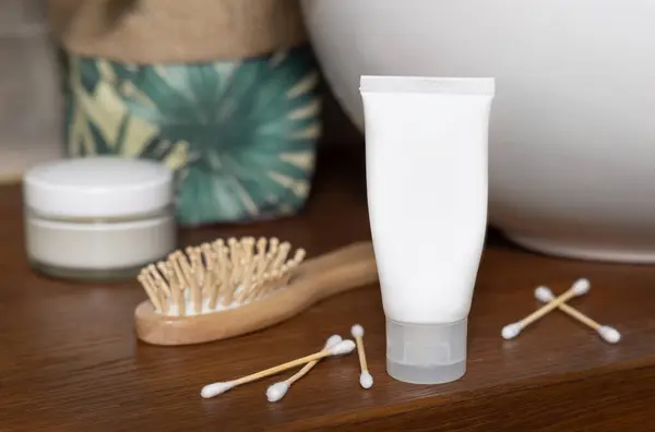 White cream tube near  bamboo hair brush, basin and cotton swabs on wooden countertop in bathroom, close up, cosmetic packaging mockup. Everyday beauty scene with sustainable products