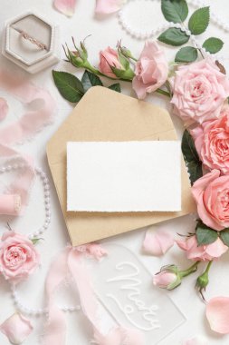 Açık pembe güllerin yanındaki boş kart ve zarf, bir kutuda nişan yüzüğü ve beyaz masa manzaralı ipek kurdeleler, düğün kırtasiyesi modeli. Pastel dekorlu romantik düz bir yatak. 