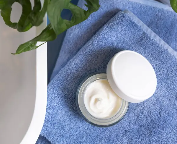 Opened cream jar with blank white lid on blue towels near basin and green monstera blue countertop, top view, mockup.  Lifestile scene with skincare beauty product in bathroom