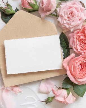 Boş kart ve zarf pembe güllerin yanında ve beyaz masa manzaralı ipek kurdeleler, düğün modeli. Yatay kart ve pastel dekoru olan romantik düz bir yatak. 