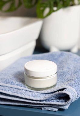 Kozmetik krem kavanozu. Mavi havluların üzerinde boş beyaz kapak ve yeşil canavar mavisi tezgah var. Yaklaş, maket yap. Tuvalette cilt bakım ürünleriyle dolu hayat dolu bir sahne.