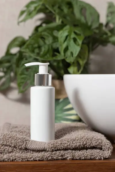 White one pump cosmetic bottle on brown folded towel near basin and green monstera on wooden countertop in bathroom close up, cosmetic mockup. Modern lifestile scene with sustainable personal care products